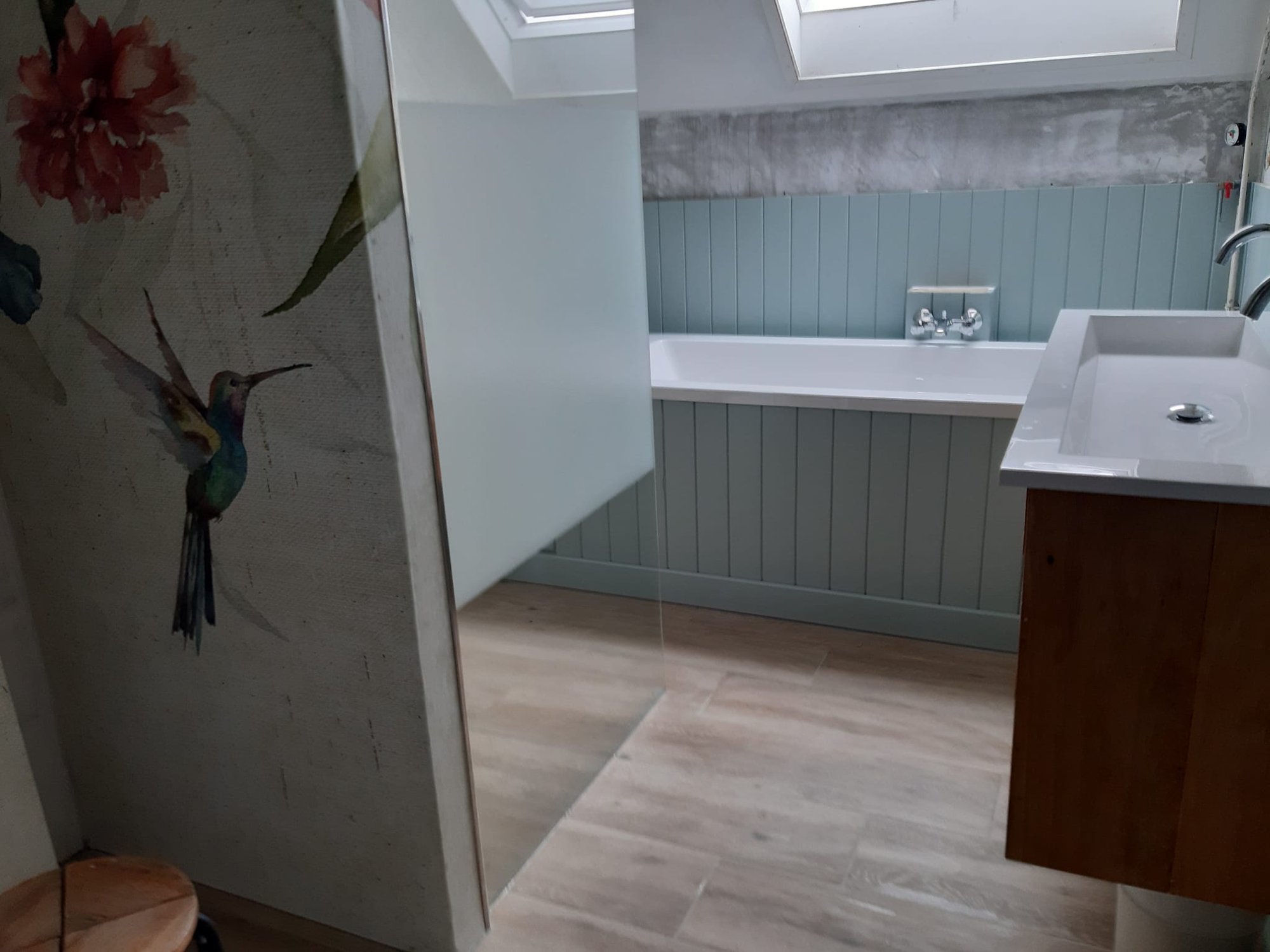 Overzicht van de badkamer met ligbad, eucalyptusgroene lambrisering, kolibrie-behang en houtlook-vloer